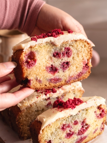 Hands holding up a slice of white chocolate raspberry loaf cake above the other slices.