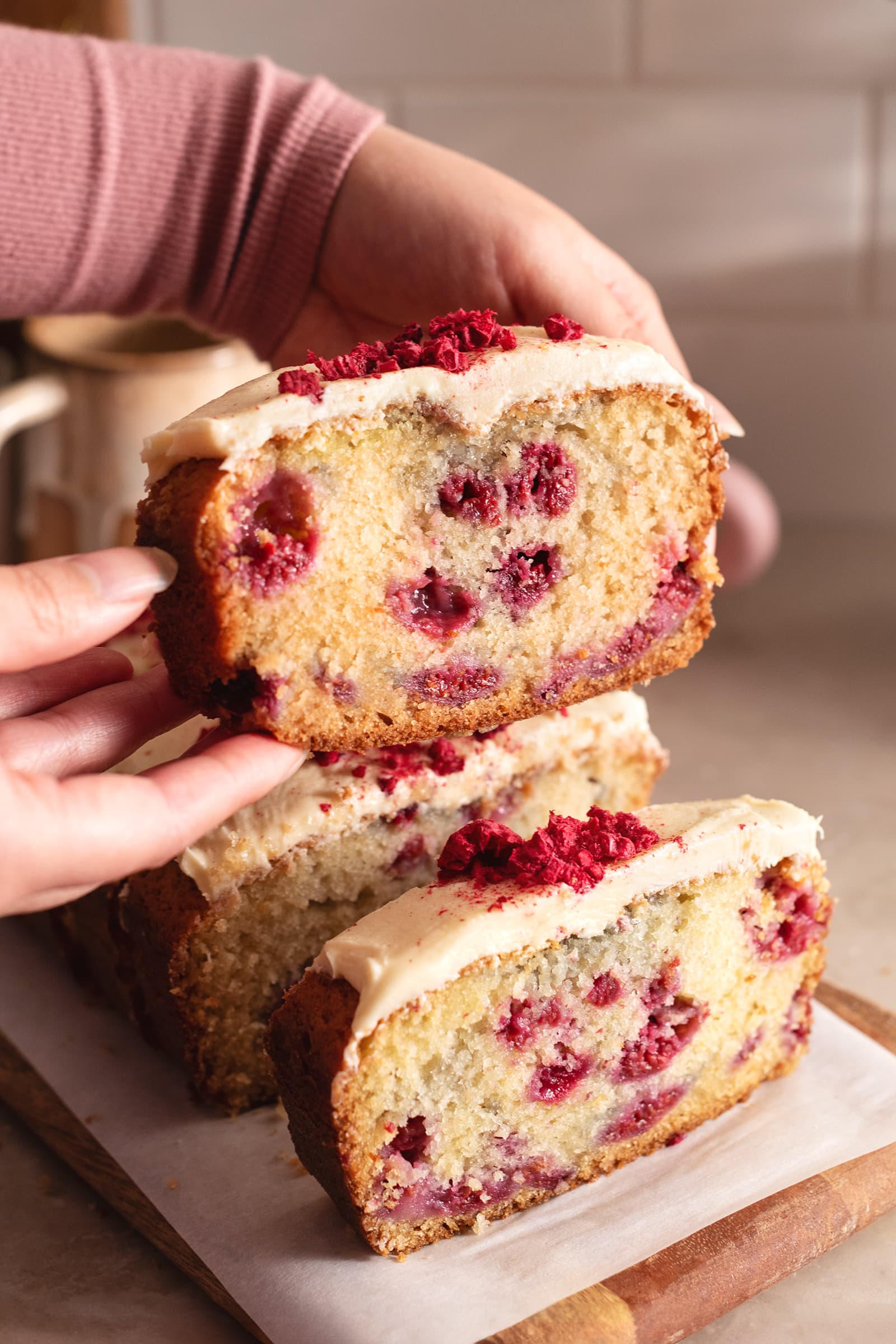 Hands holding up a slice of white chocolate raspberry loaf cake above the other slices.