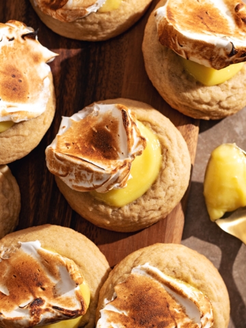 Several lemon meringue cookies scattered on a wooden board next to a spoonful of lemon curd.