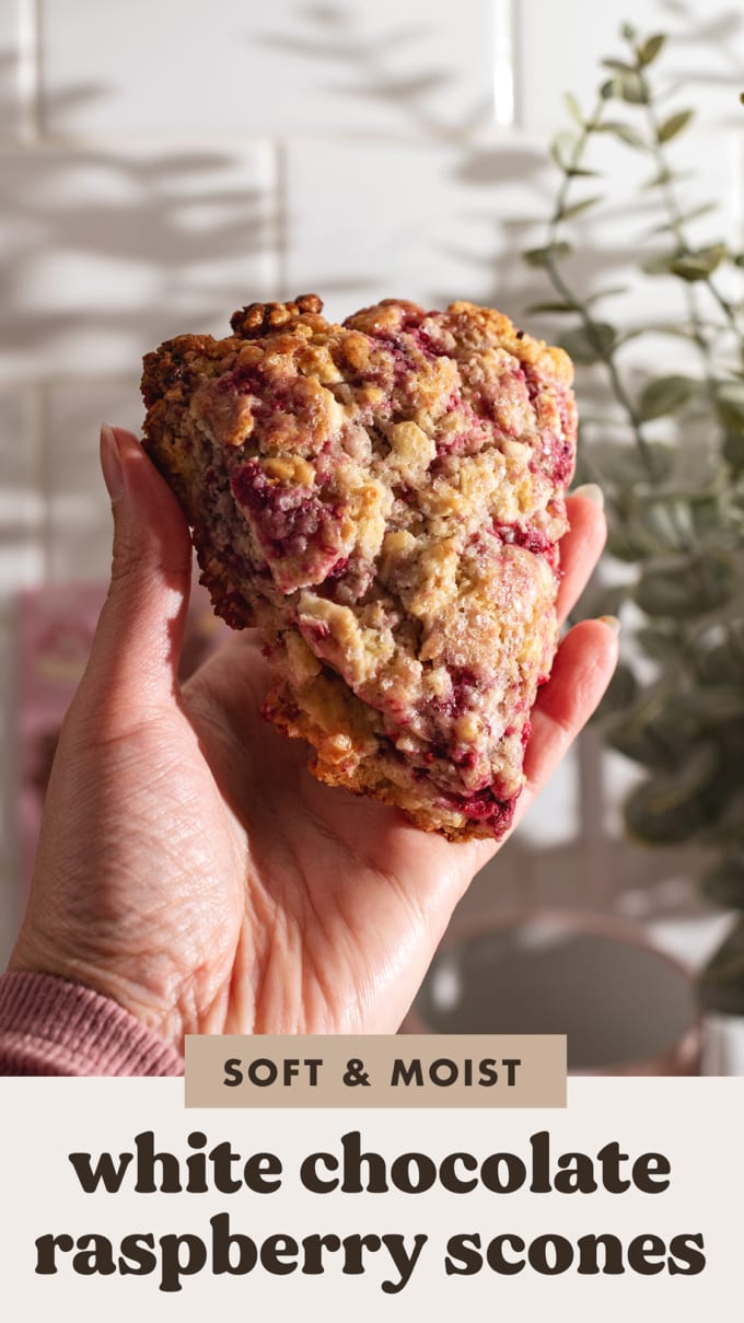 A hand holding up a baked white chocolate raspberry scone in front of a kitchen wall.