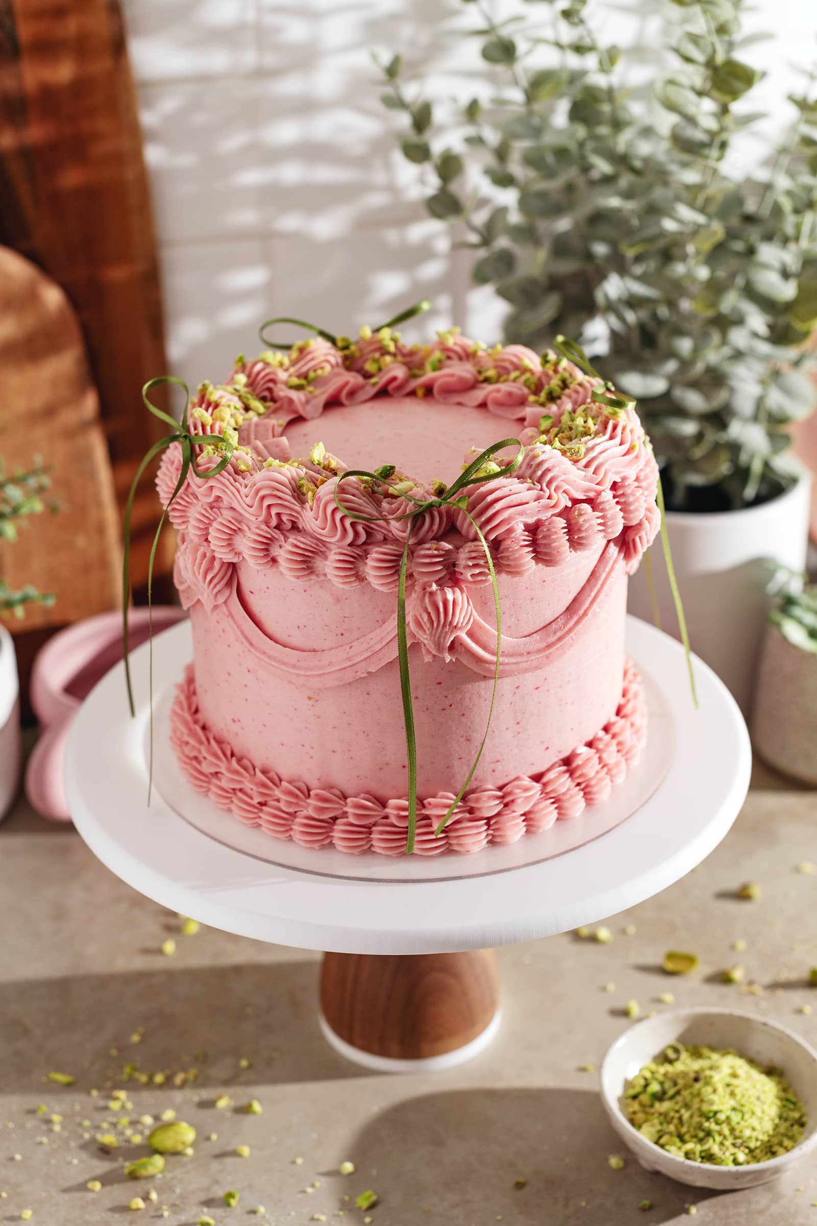 A raspberry pistachio cake on a cake stand covered with an elegant buttercream design and decorated with ribbon bows.