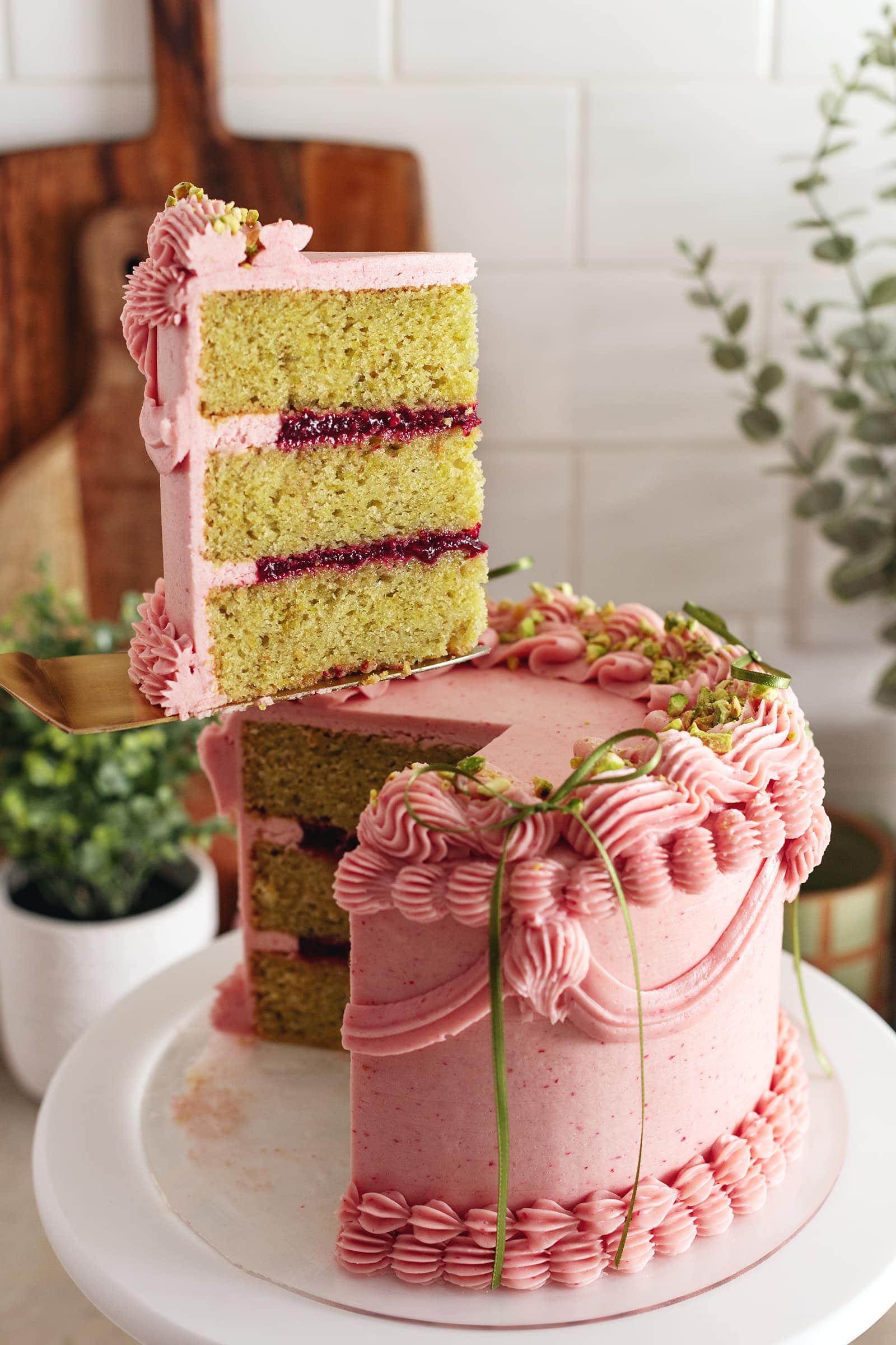 A slice of raspberry pistachio cake lifted above the rest of the cake with a cake server.