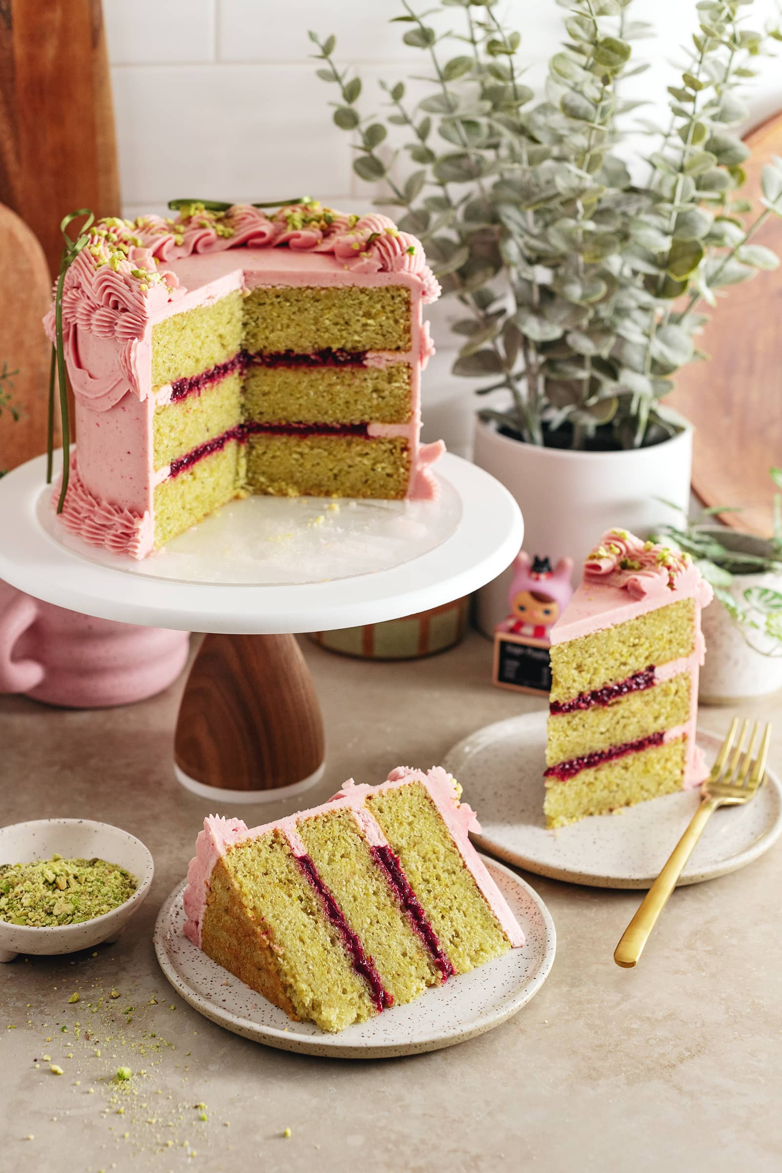 A raspberry pistachio cake on a cake stand with slices of cake on plates around it.