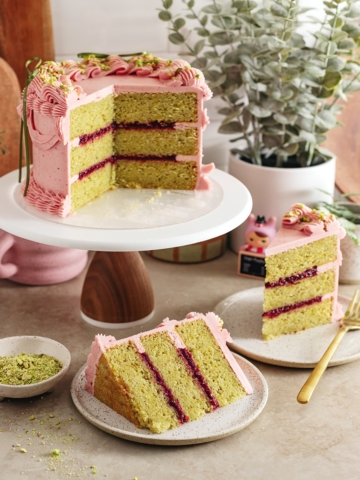 A raspberry pistachio cake on a cake stand with slices of cake on plates around it.