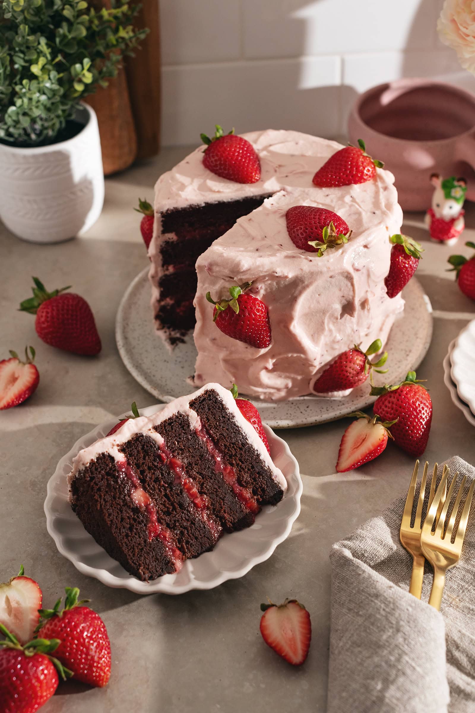 A slice of strawberry chocolate cake on a plate in front of the rest of the strawberry-covered cake.