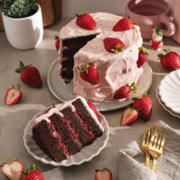 A slice of strawberry chocolate cake on a plate in front of the rest of the strawberry-covered cake.