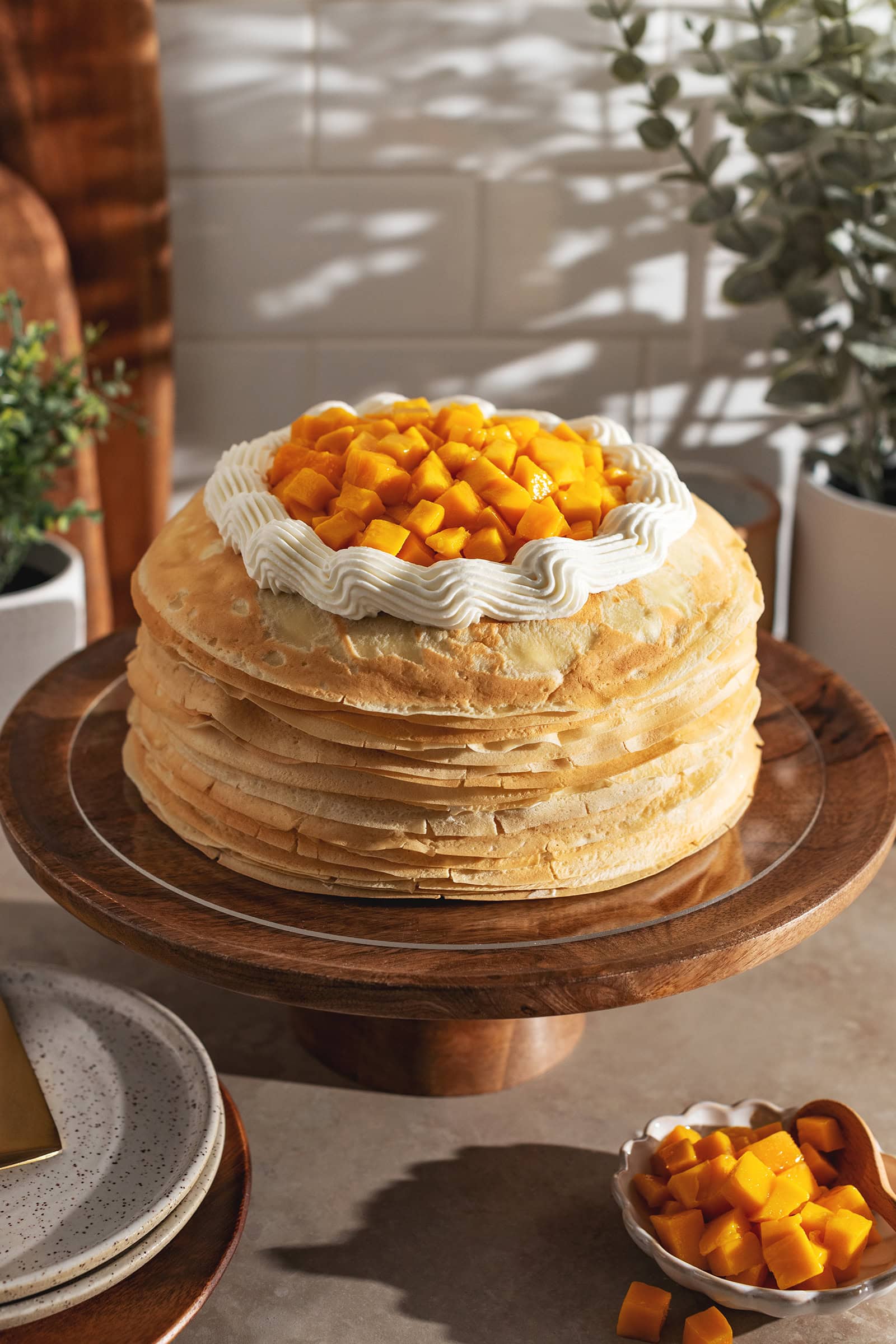 A mango crepe cake on a wooden cake stand topped with a ring of whipped cream and fresh mango chunks.