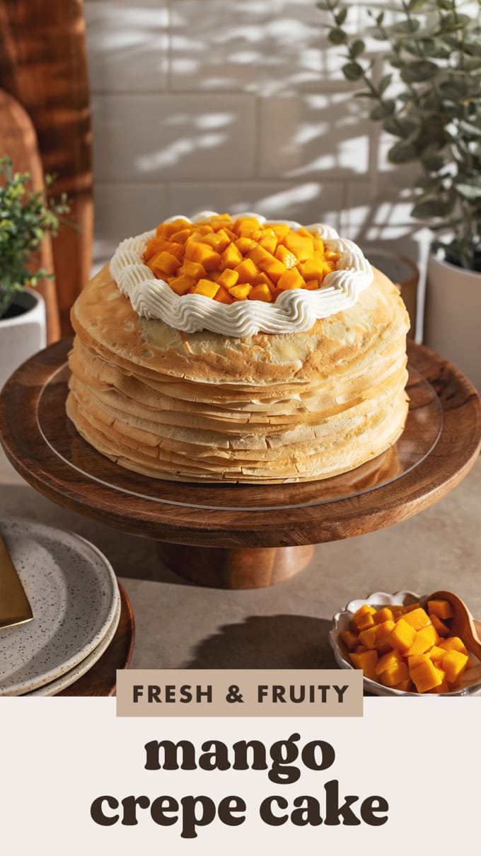 A mango crepe cake on a wooden cake stand topped with a ring of whipped cream and fresh mango chunks.