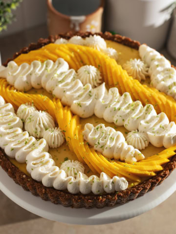 A mango tart decorated with squiggles of whipped cream and fresh mangoes on a cake stand.