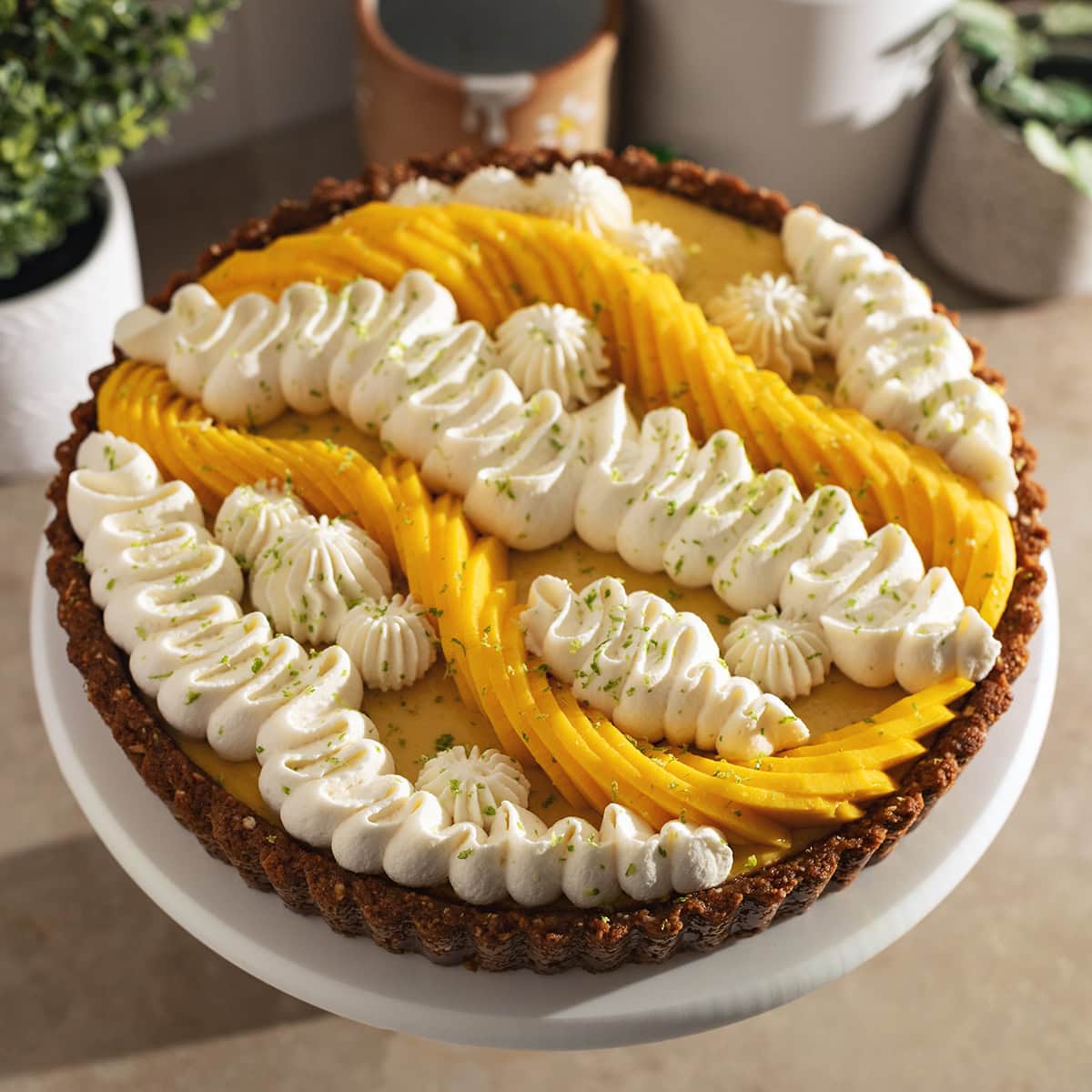 A mango tart decorated with squiggles of whipped cream and fresh mangoes on a cake stand.