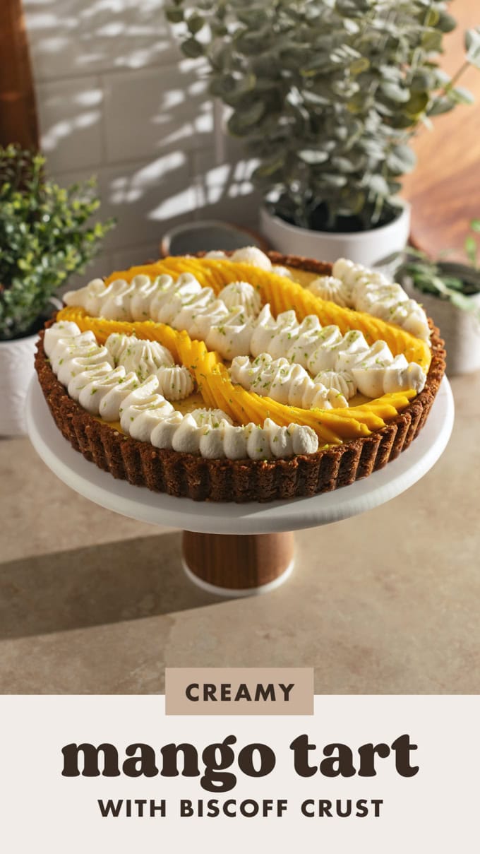 A mango tart decorated with squiggles of whipped cream on a cake stand on a kitchen counter.