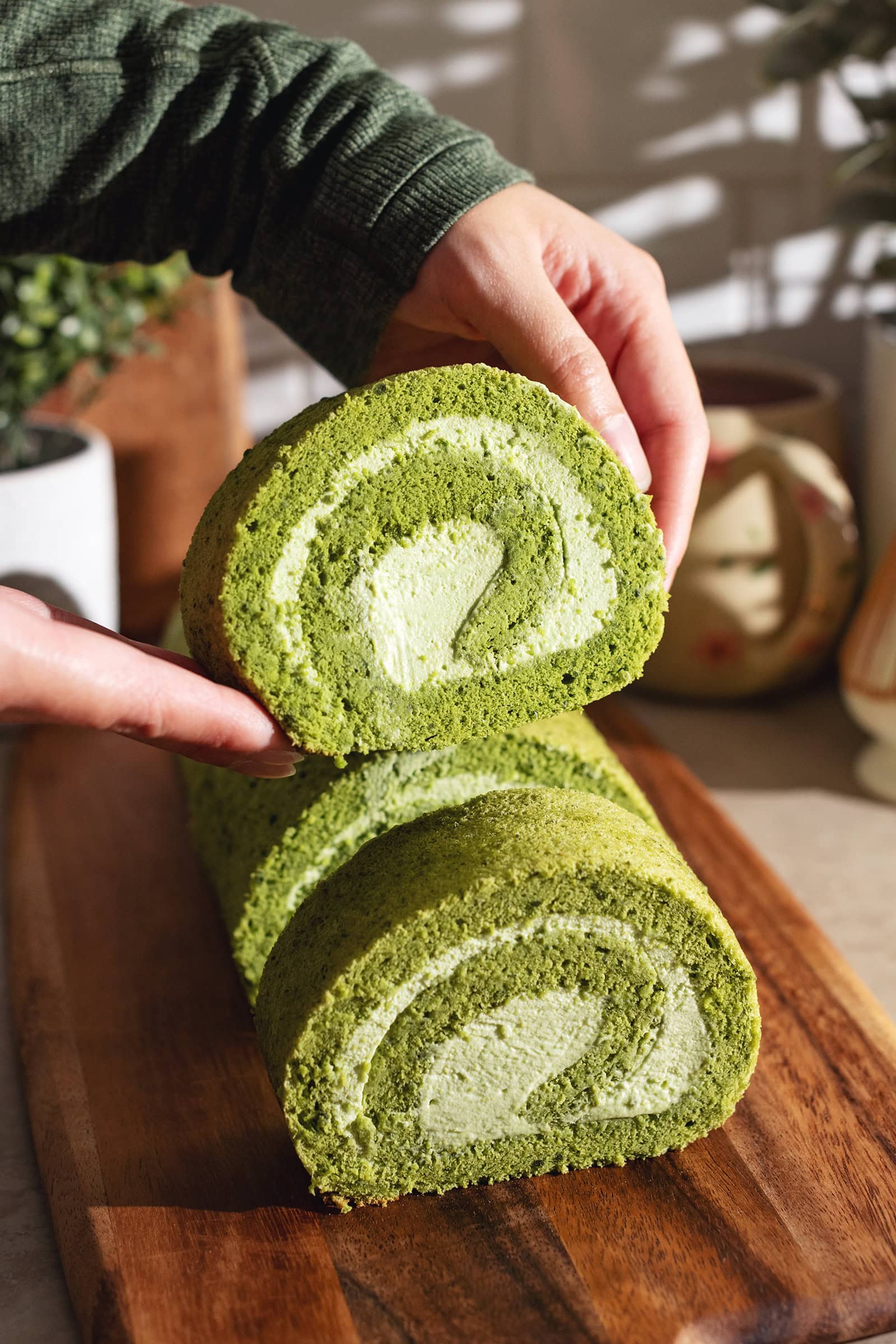 Hands holding a slice of matcha roll cake above the rest of the cake.