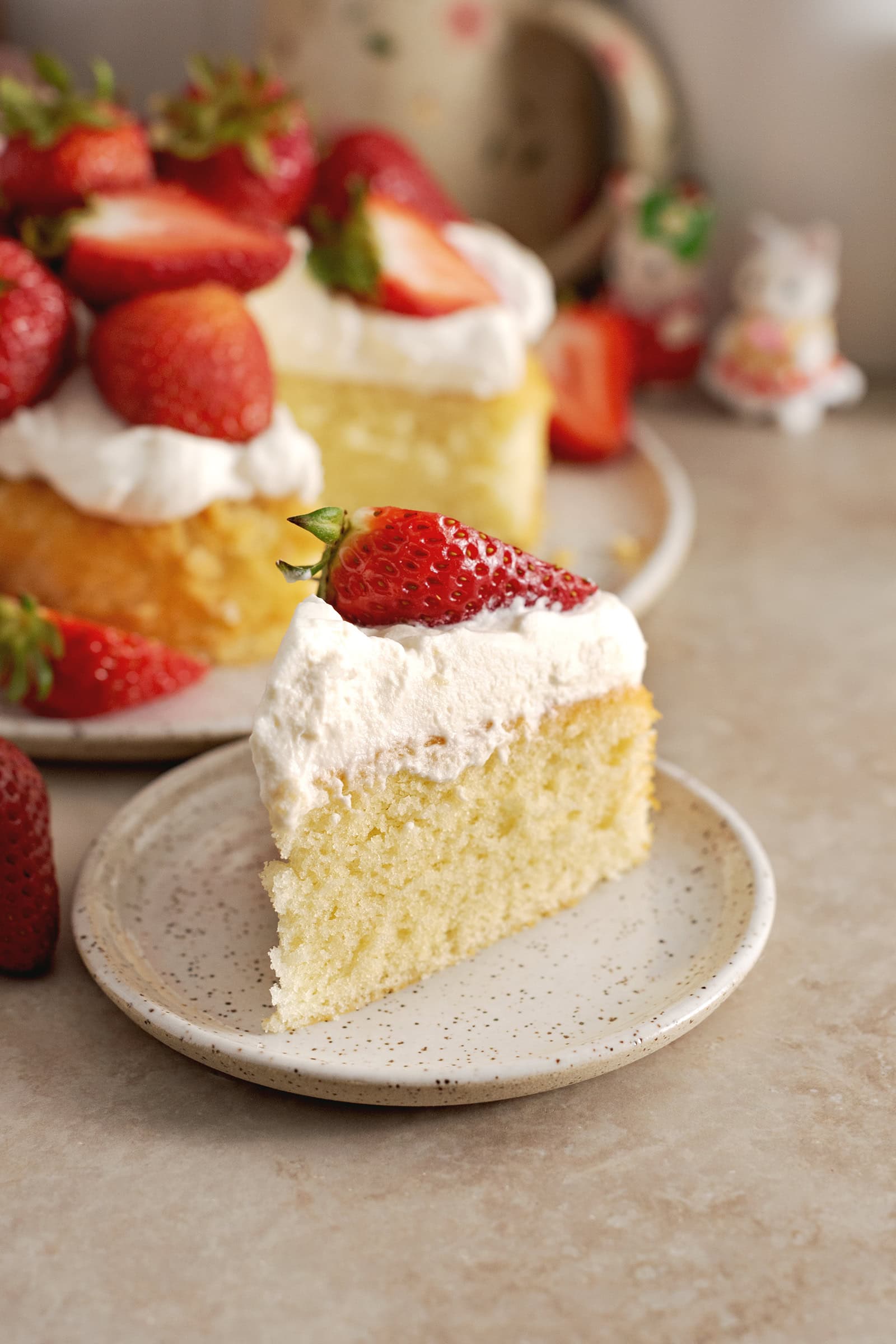 A slice of strawberry cream cake on a plate in front of the rest of the cake.