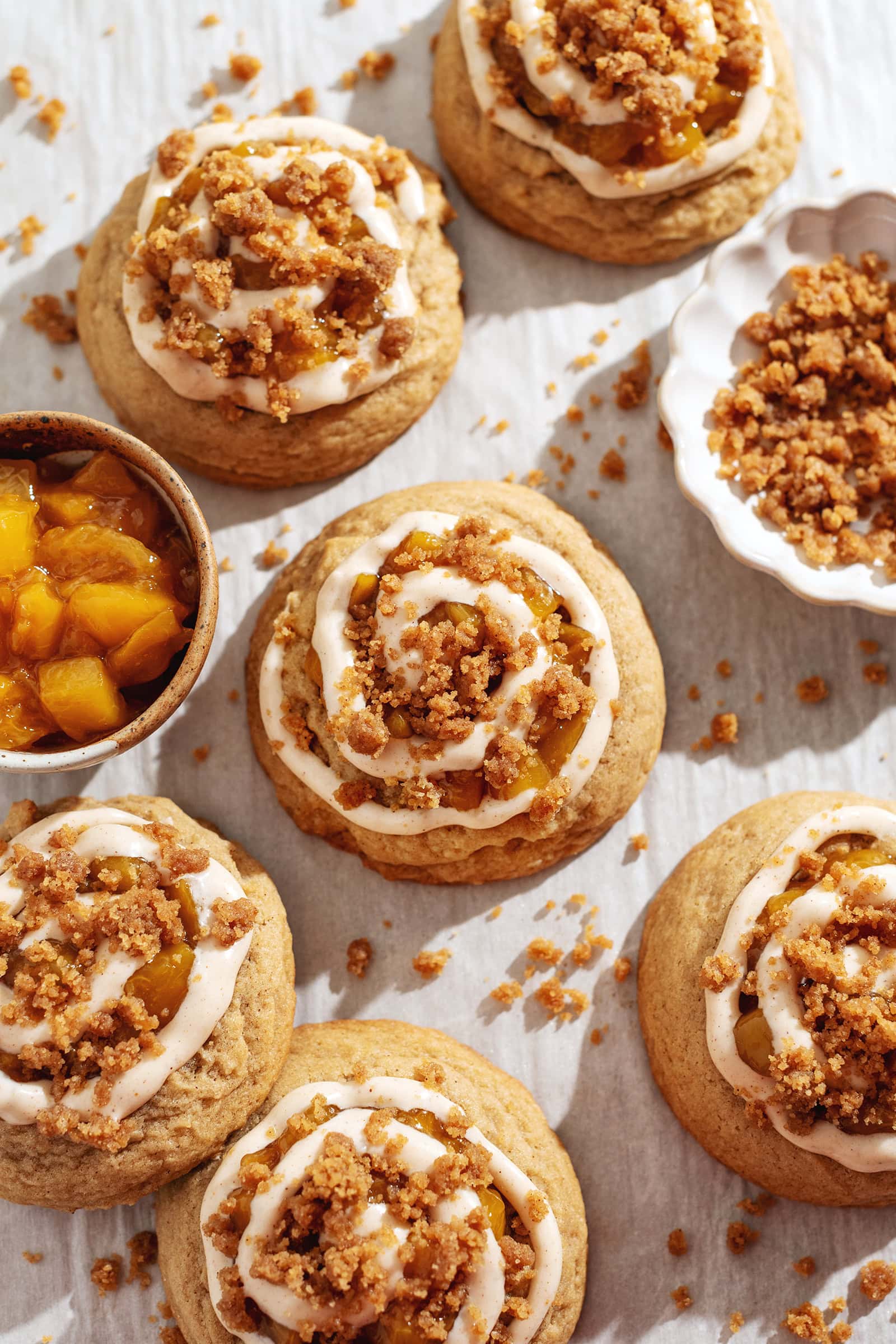 Several peach cobbler cookies scattered on parchment paper surrounded by baked crumble and a bowl of peach compote.