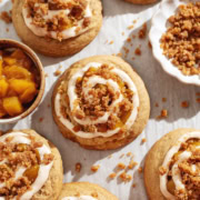 Several peach cobbler cookies scattered on parchment paper surrounded by baked crumble and a bowl of peach compote.