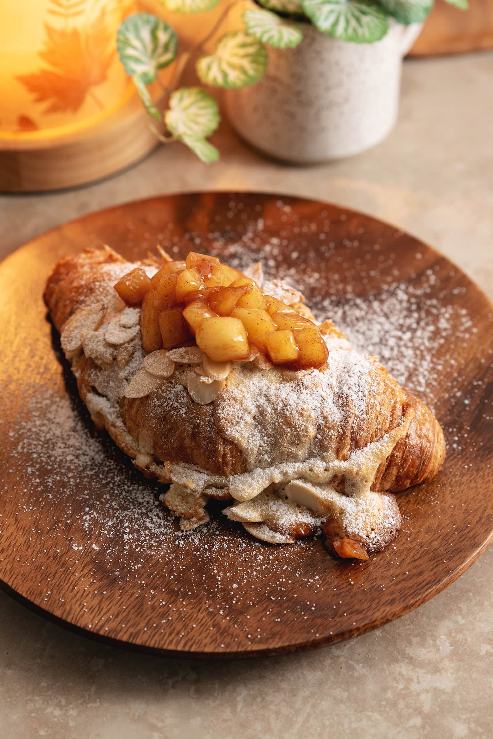 An apple pie almond croissant dusted with powdered sugar on a wooden plate.