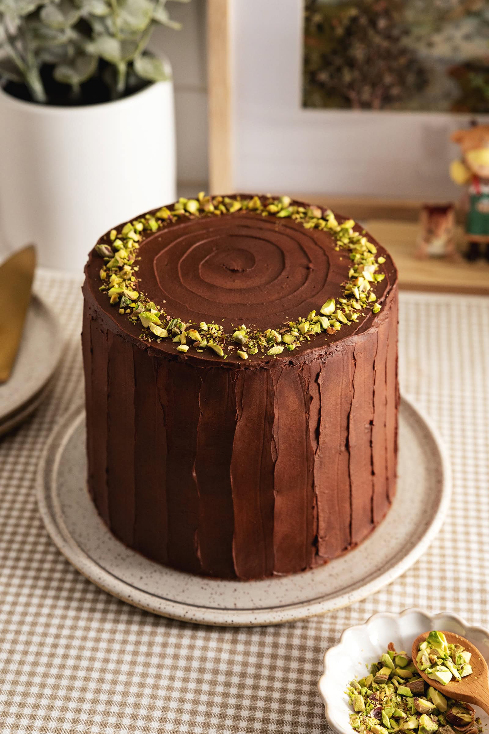 A Dubai chocolate pistachio cake frosted with chocolate ganache and topped with a ring of pistachios on a plate.