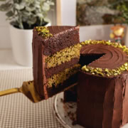 A slice of Dubai chocolate pistachio cake being lifted above the rest of the cake with a cake server.