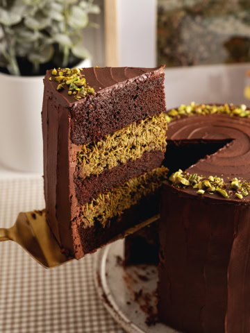 A slice of Dubai chocolate pistachio cake being lifted above the rest of the cake with a cake server.
