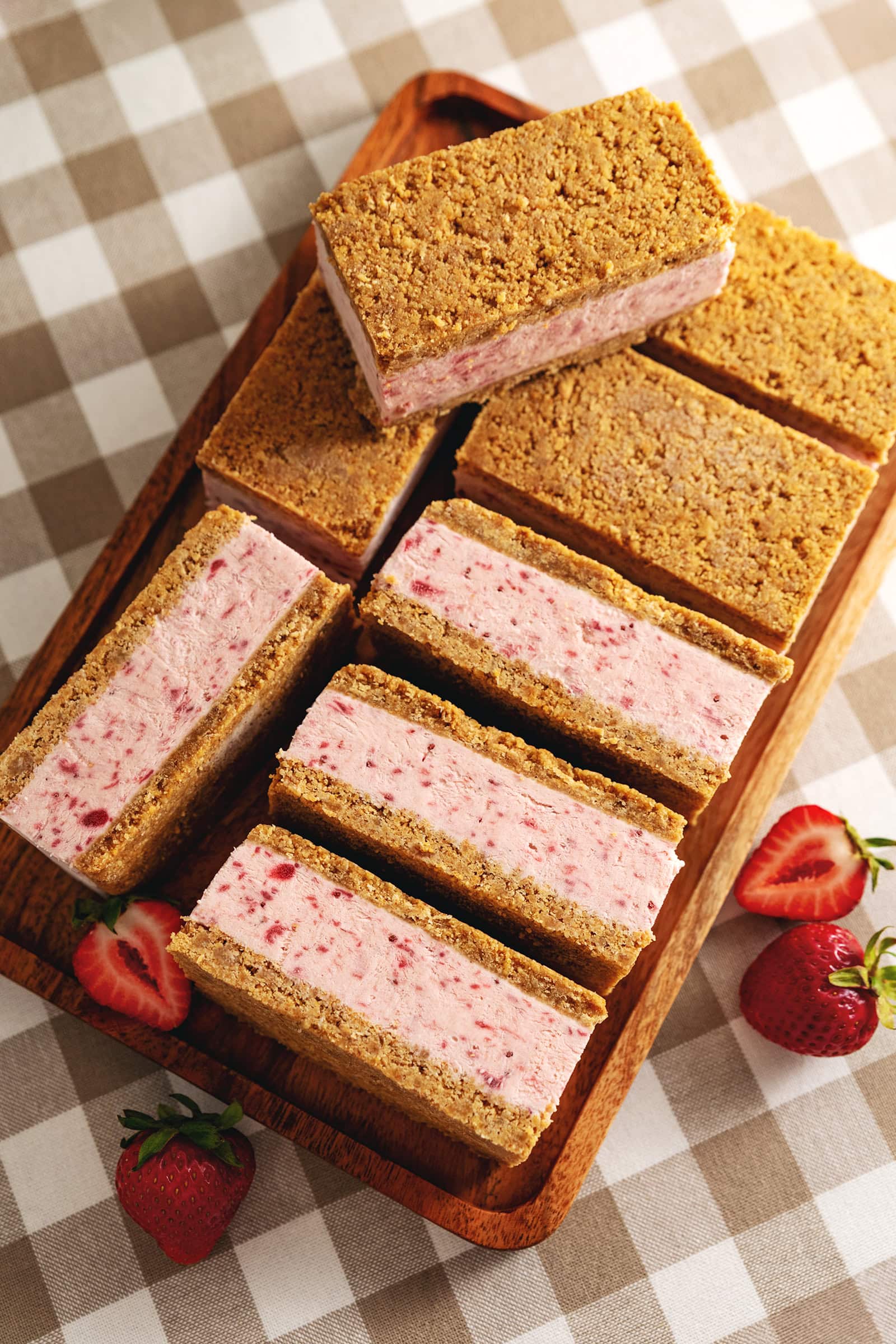 Strawberry ice cream sandwiches lined up on a wooden tray with fresh strawberries around it.