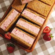 Strawberry ice cream sandwiches lined up on a wooden tray with fresh strawberries around it.