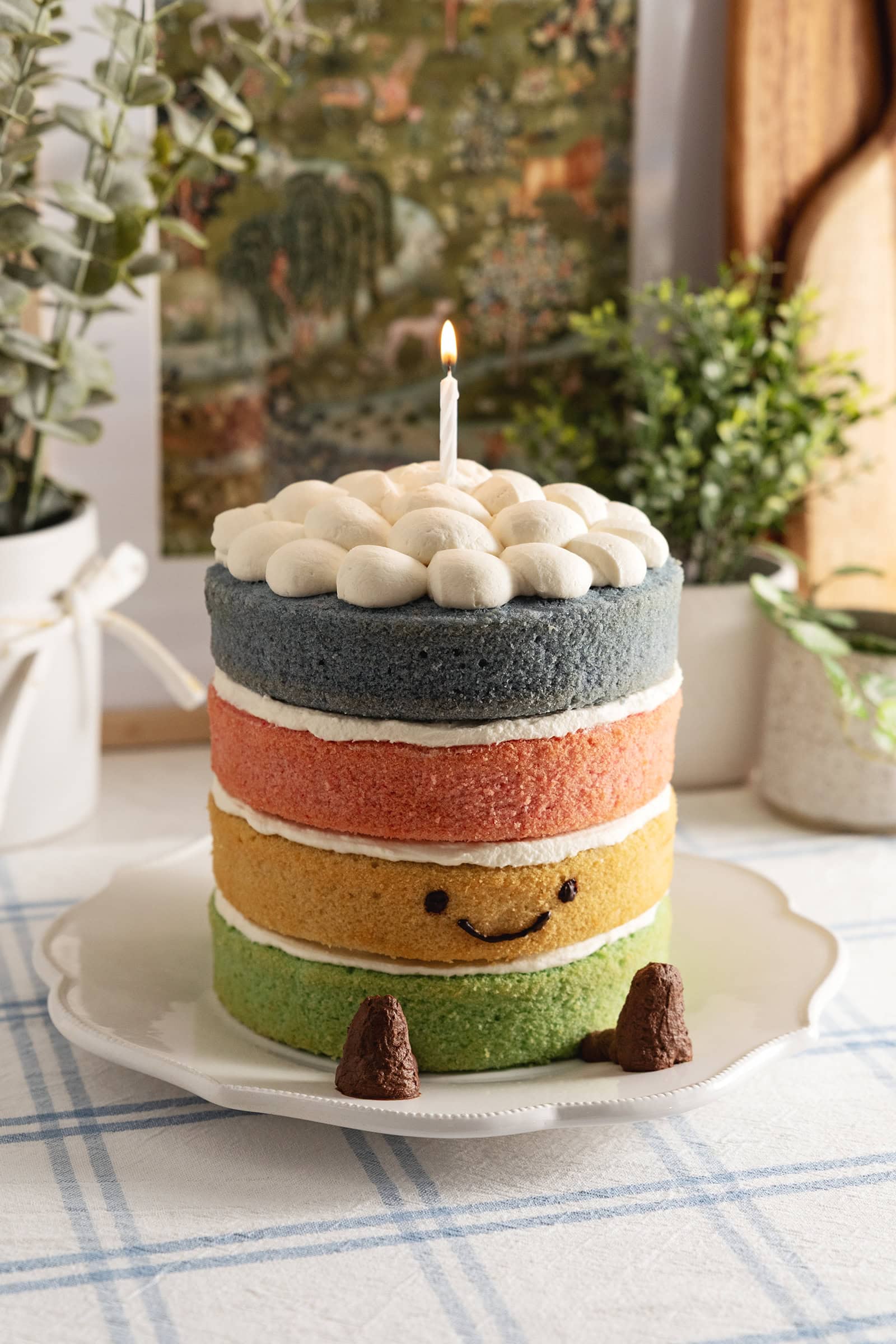 A 4-layer Jellycat rainbow cake with a smiley face, legs, and a lit candle on top on a kitchen counter.