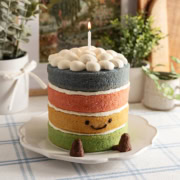 A 4-layer Jellycat rainbow cake with a smiley face, legs, and a lit candle on top on a kitchen counter.