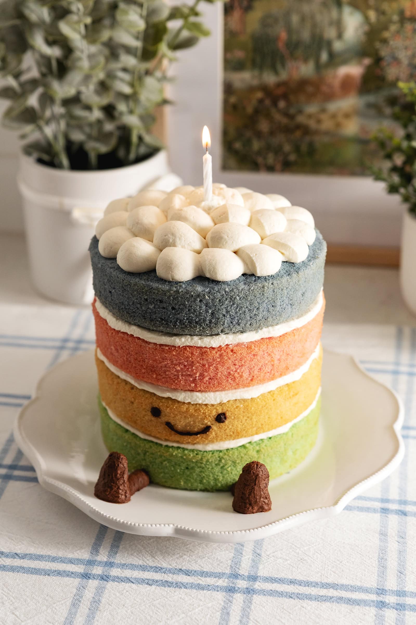 A 4-layer Jellycat rainbow cake with a smiley face, legs, and a lit candle on top on a kitchen counter.