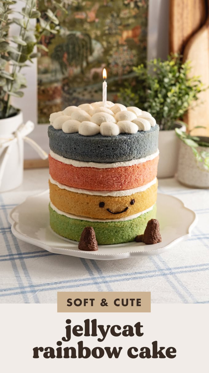 A 4-layer Jellycat rainbow cake with a smiley face, legs, and a lit candle on top on a kitchen counter.