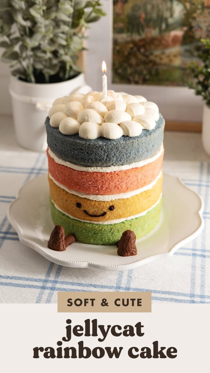 A 4-layer Jellycat rainbow cake with a smiley face, legs, and a lit candle on top on a kitchen counter.