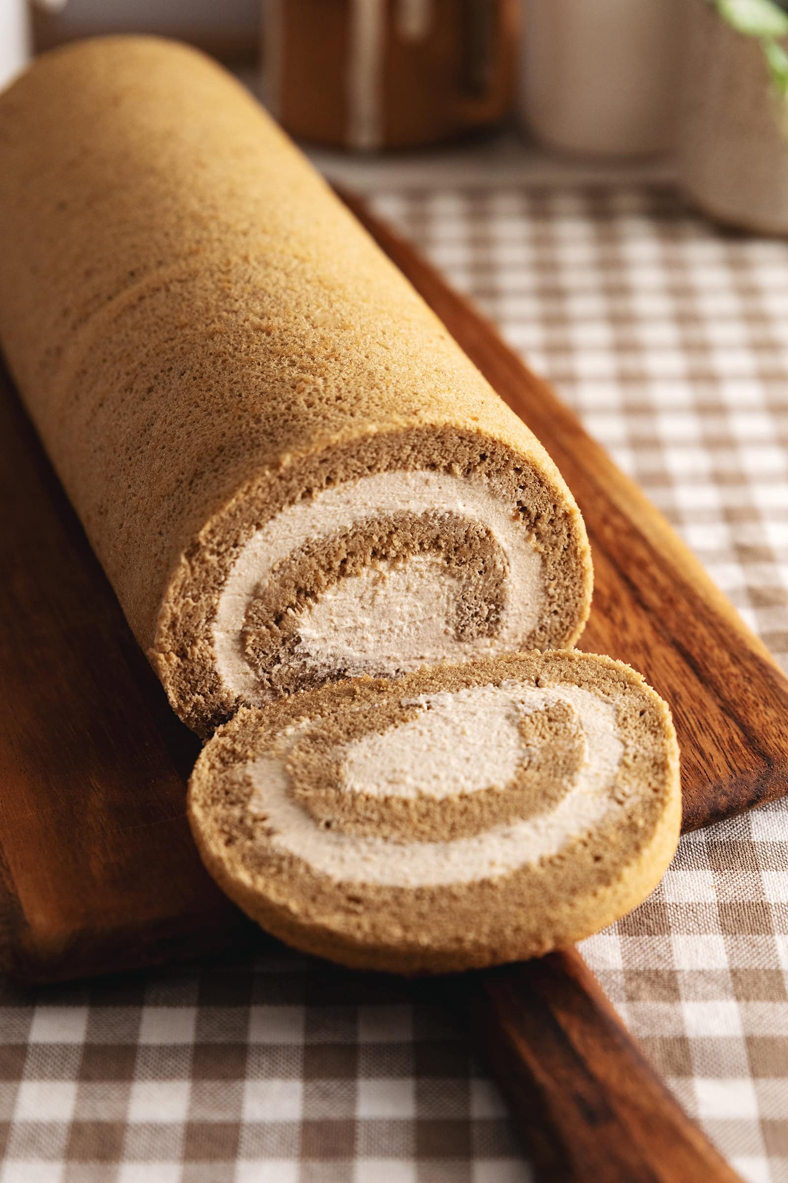 A coffee swiss roll on a wooden board with one slice cut and laying down in front of the rest of the cake.