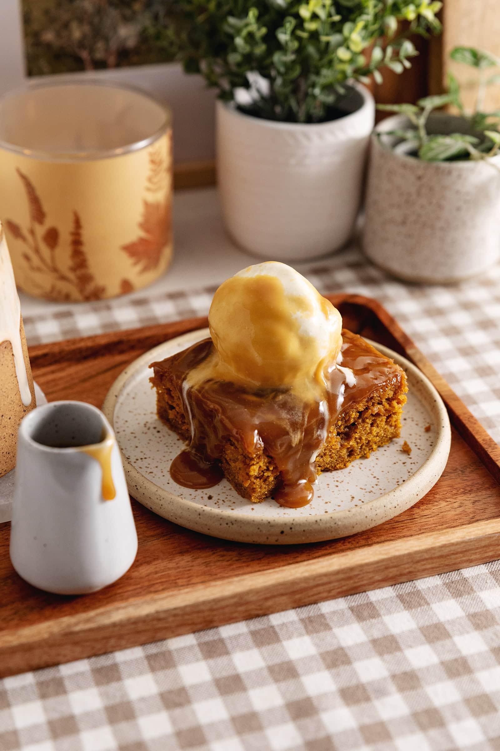 A slice of pumpkin sticky toffee pudding on a wooden tray with a small cup of toffee sauce next to it.