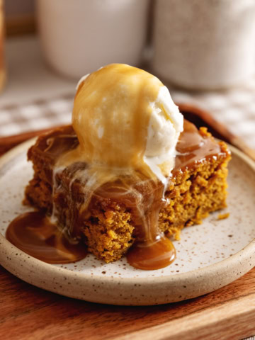 A slice of pumpkin sticky toffee pudding on a plate with a scoop of ice cream on top and toffee sauce dripping down.