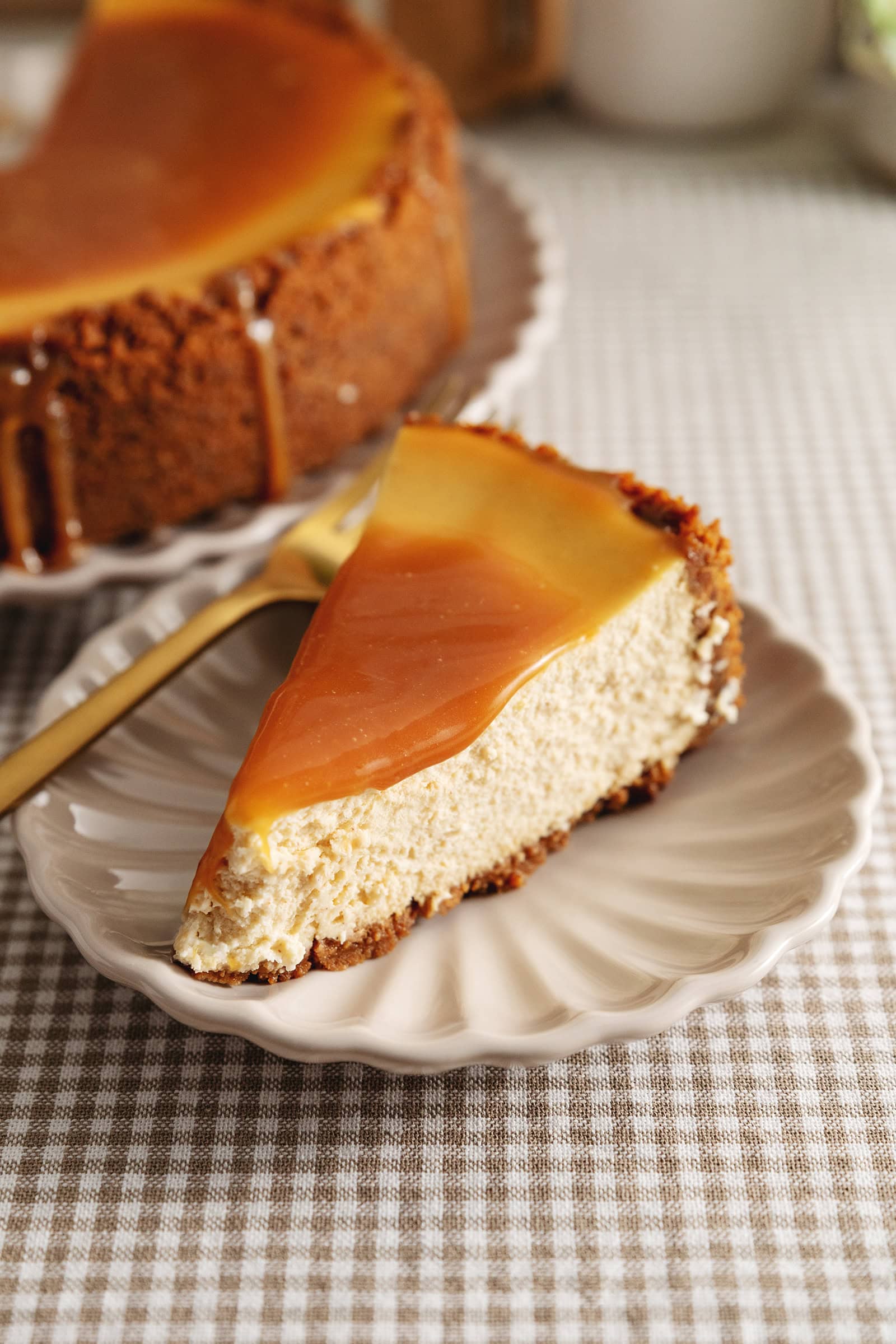 A slice of salted caramel cheesecake on a plate in front of the rest of the cheesecake.