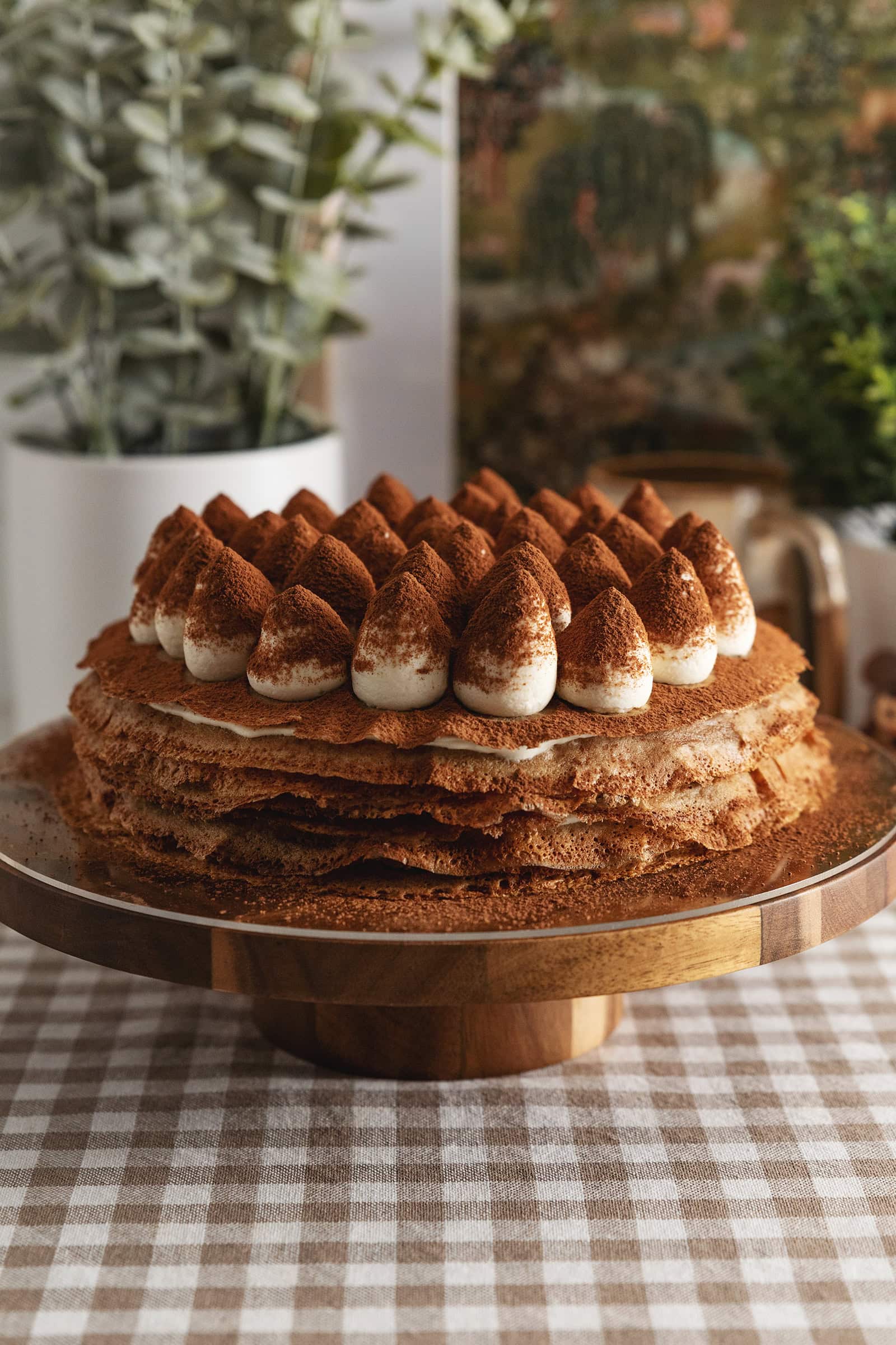 A tiramisu crepe cake on a wooden cake stand with dollops of mascarpone cream on top dusted with cocoa powder.