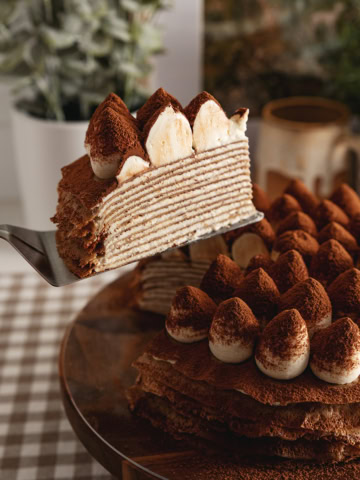 A slice of tiramisu crepe cake being lifted with a cake server from the rest of the cake to show the crepe layers inside.