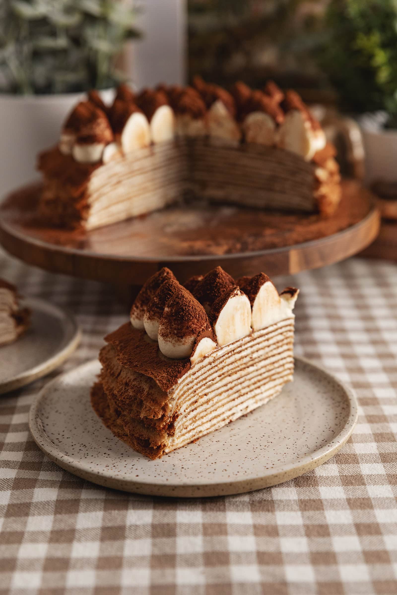 A slice of tiramisu crepe cake on a plate with the rest of the cake on a cake stand in the background.