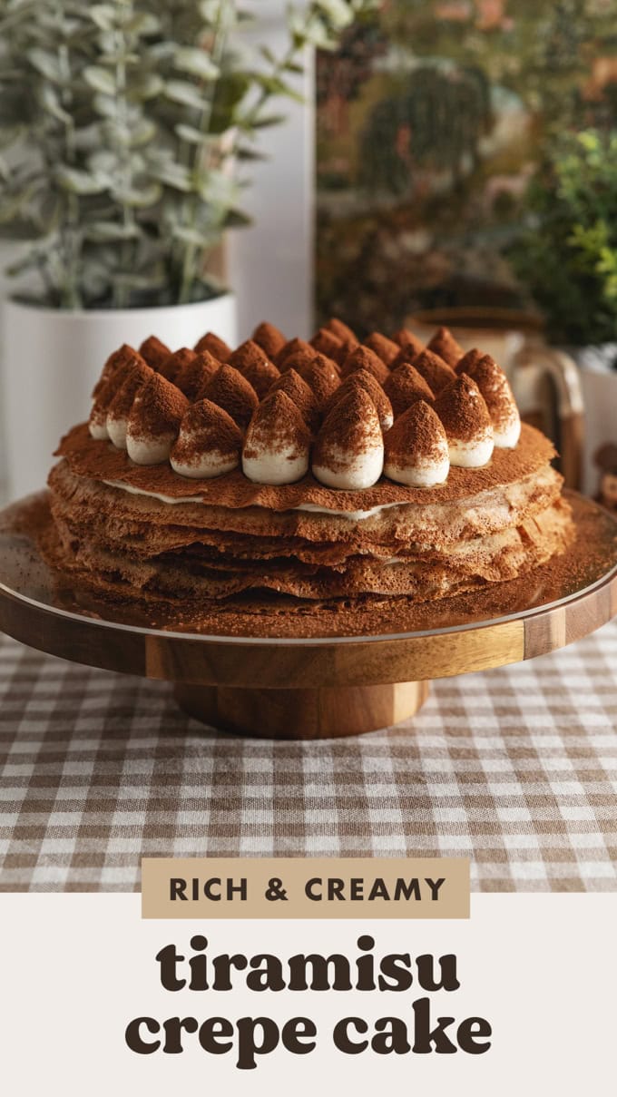 A tiramisu crepe cake on a wooden cake stand with dollops of mascarpone cream on top dusted with cocoa powder.