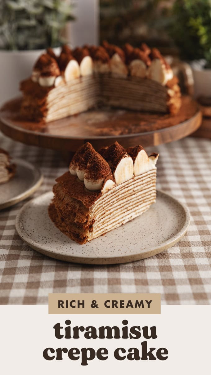 A slice of tiramisu crepe cake on a plate with the rest of the cake on a cake stand in the background.