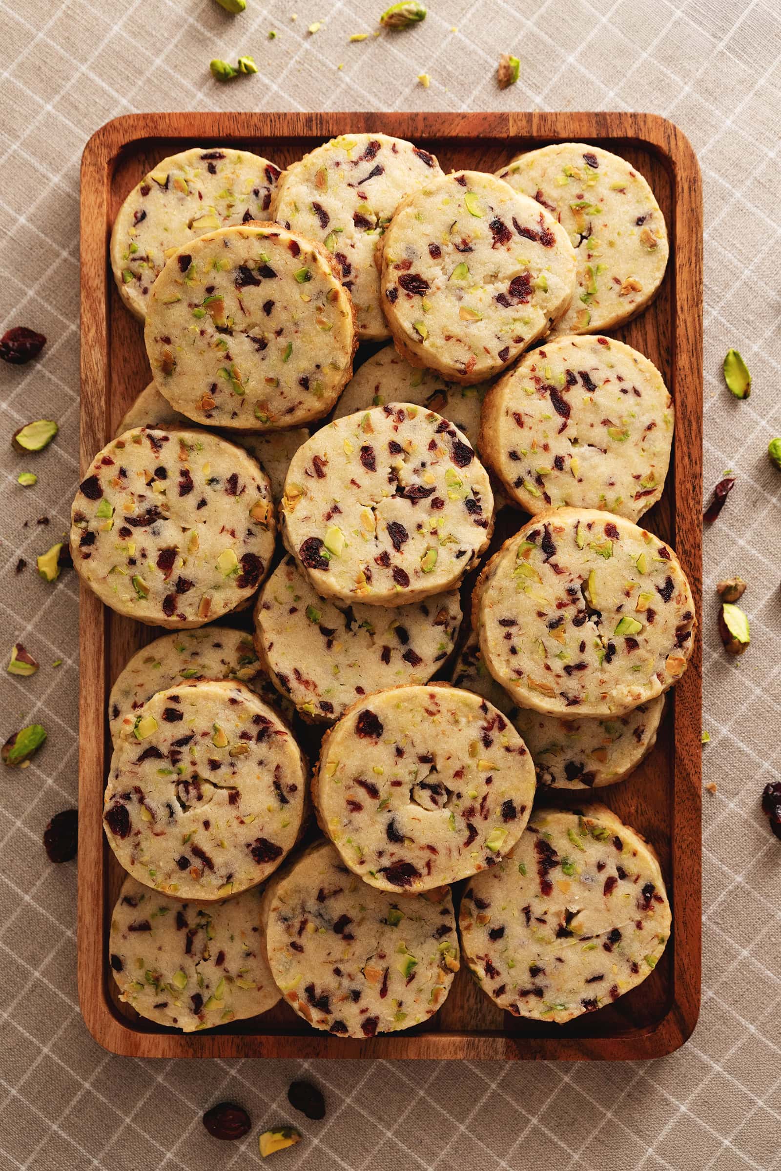 A wooden platter full of cranberry pistachio shortbread cookies stacked on top of each other.