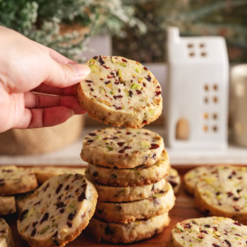 A hand holding a cranberry pistachio shortbread cookies on top of a stack of cookies.