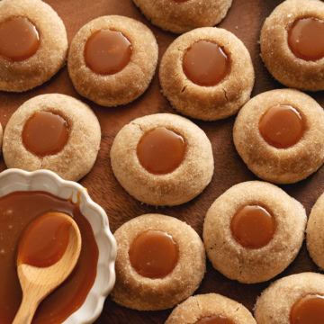 Several gingerbread thumbprint cookies scattered on a wood tray with a dish of caramel to the side.